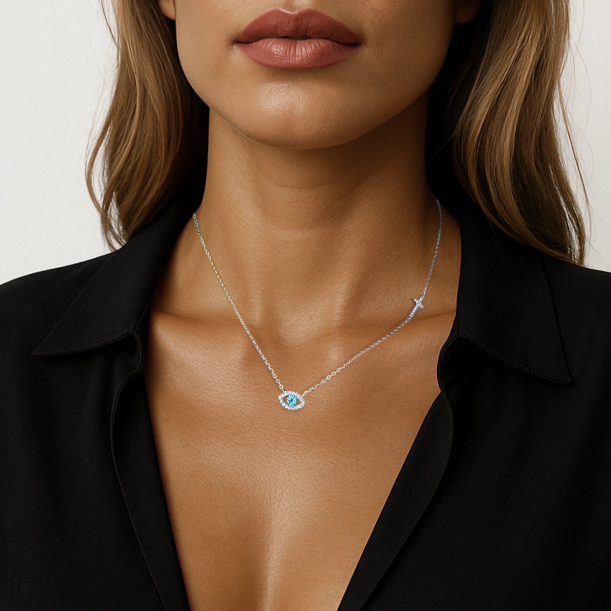Close-up of a woman wearing a sterling silver necklace featuring a dainty evil eye pendant with a sparkling blue center and a side-set cross charm, styled with a deep V-neck black blouse.