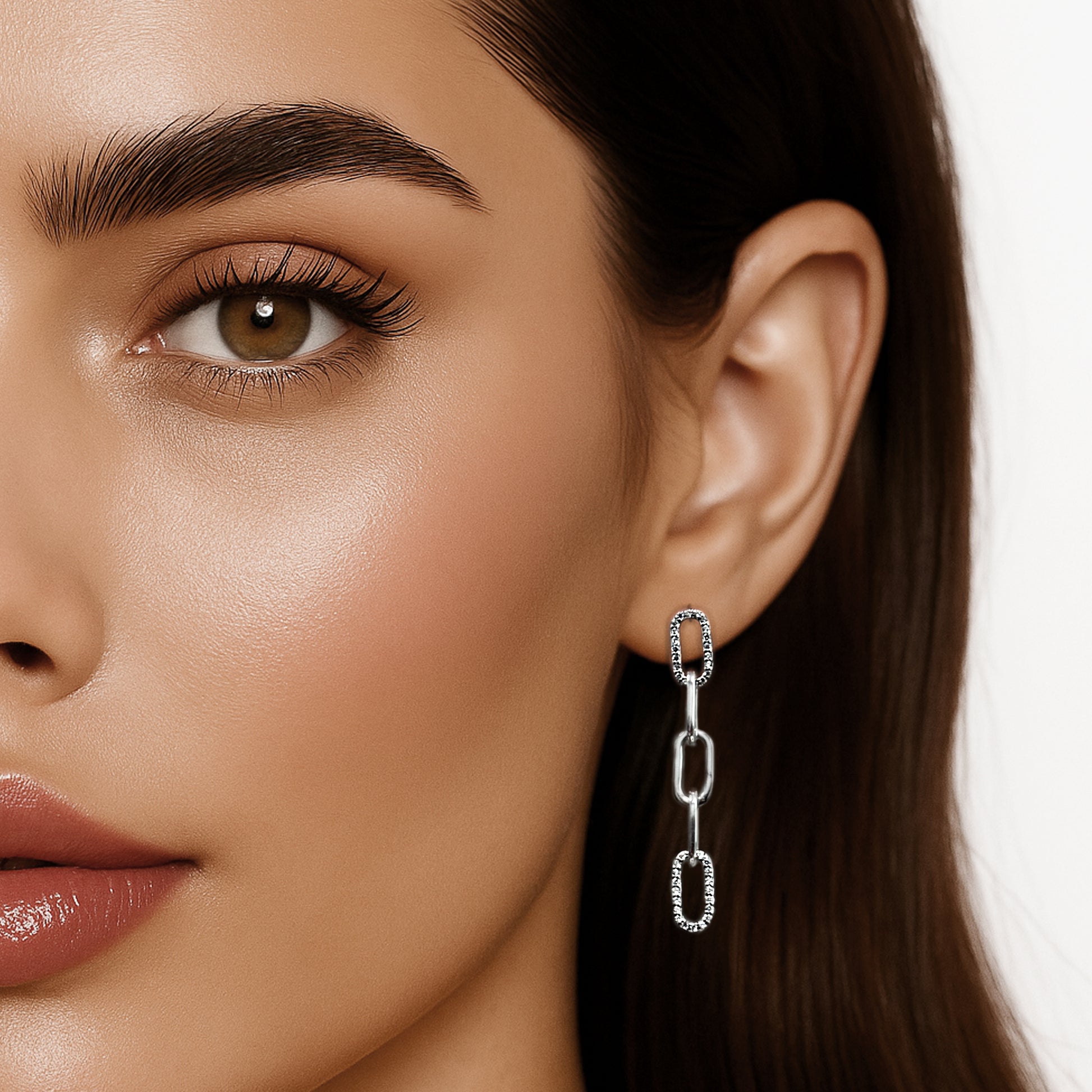 Extreme close-up of a woman’s half face showcasing a sparkling asymmetric chain drop earring with cubic zirconia links. Her glossy lips, hazel eye, and centered ear are highlighted against a clean white background, with soft lighting enhancing the polished silver finish.