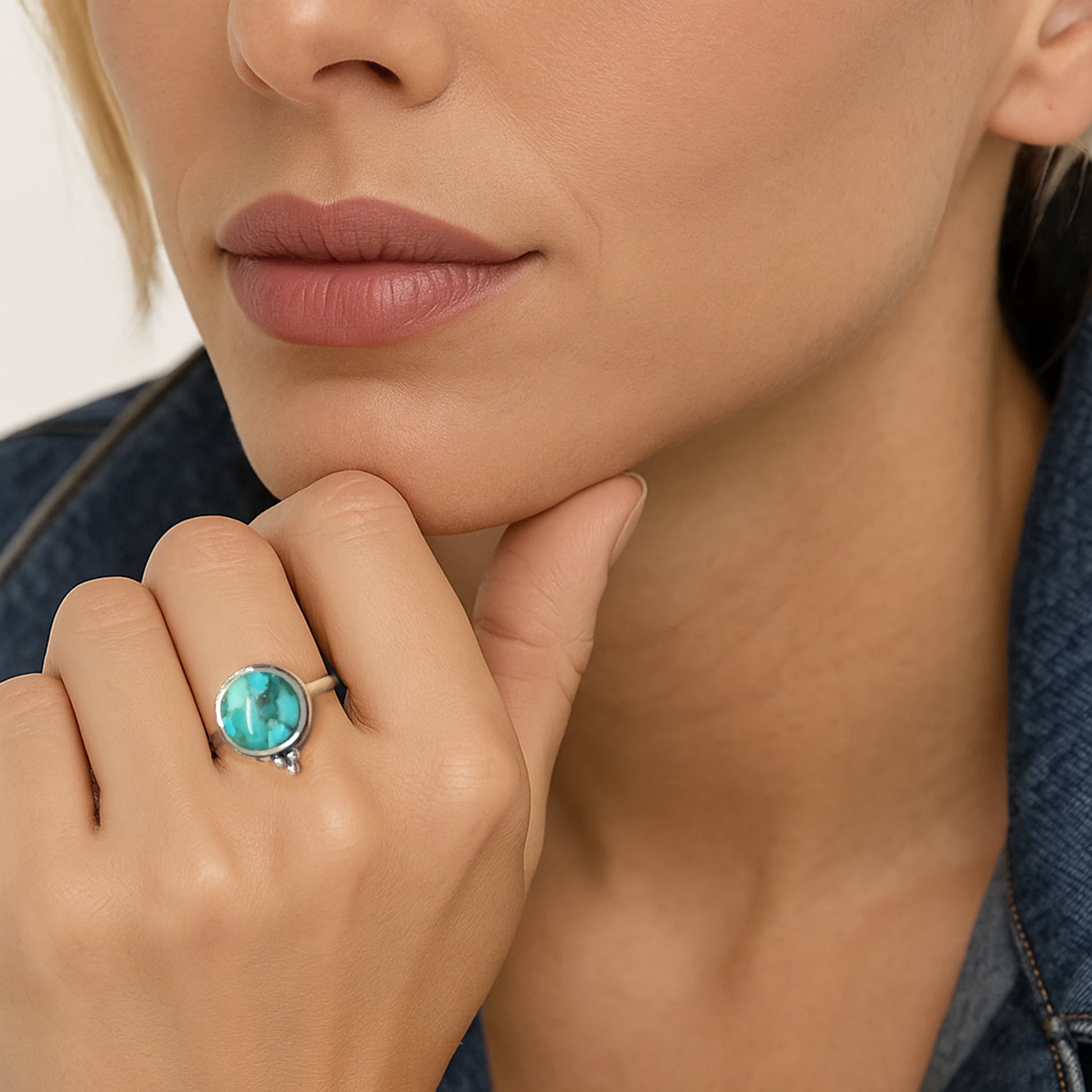 Close-up image of a woman's hand adorned with a bold silver ring featuring a round turquoise stone. The ring stands out against her natural-toned makeup and denim jacket, with her hand gently resting on her chin in a poised, thoughtful pose.