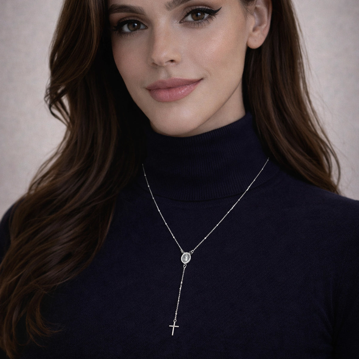 Model wearing a minimalist sterling silver rosary necklace with Miraculous Medal and cross charm over a navy blue turtleneck.