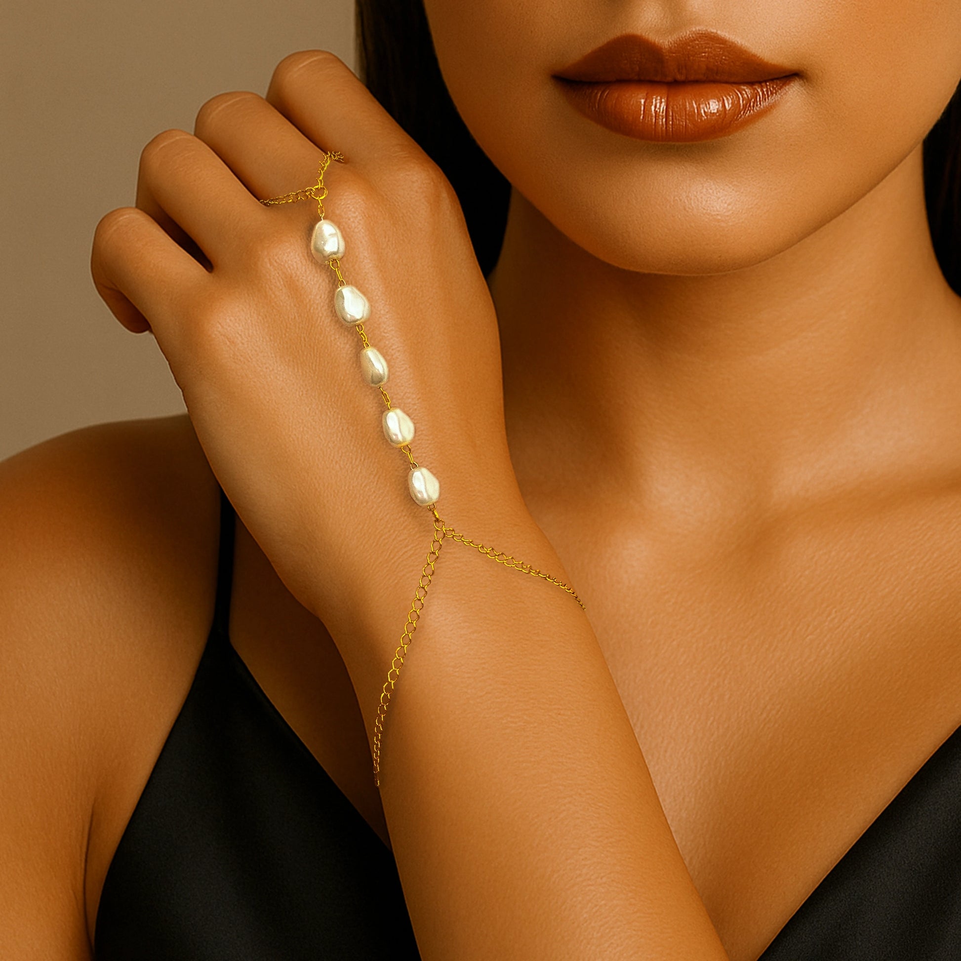 Model wearing an 18K gold filled hand harness bracelet with imitation baroque pearls, elegantly draped across the back of the hand, styled with a black satin outfit and warm bronze makeup.