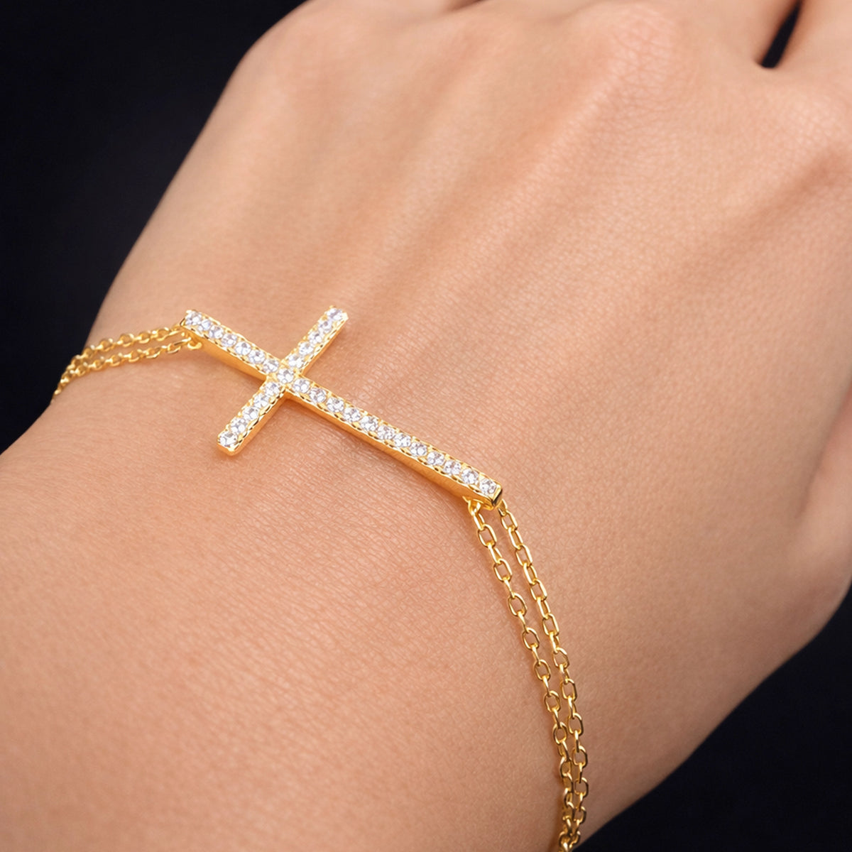 Gold cross bracelet with sparkling CZ stones on double gold chain, worn on wrist, close-up studio photo with soft neutral background, elegant Christian jewelry from 108 Creations